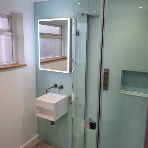 Modern bathroom, a shower enclosure on the right and a cube shaped sink and led lit mirror the the left of the shower. Light wood floor and turquoise walls