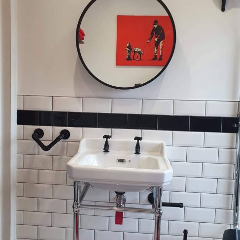 Industrial type sink area, in a bathroom. Black pipe towel rail white sink, chrome pipe sin stand, black and white tile splashback and a round mirror, above the sink