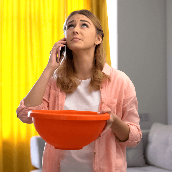 A lady, holding an orange bowl, to catch water dripping from the ceiling, whilst holding a phone in the other hand